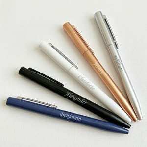 May include: Five personalized pens in various colors: navy blue, black, white, rose gold, and silver. Each pen has a name engraved on the side: Benjamin, Alexander, Charlotte, Henry, and William. The pens are arranged on a white surface.