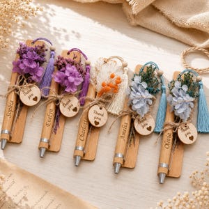 May include: Five bamboo pens attached to rectangular wooden bookmarks, each personalized with a name and decorated with dried flowers, a matching tassel, and a small circular wooden tag engraved with the text Thank you and a heart symbol.