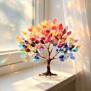 Op de afbeelding: Een decoratieve boomsculptuur met een heldere basis en een bruine stam. De bladeren zijn een levendige mix van rood, oranje, geel, groen, blauw en paars. De boom staat op een vensterbank, met zonlicht dat een regenboogeffect cre&euml;ert.