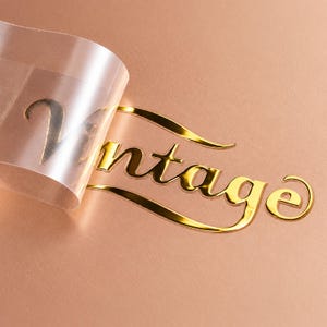May include: A gold metallic sticker featuring the word Vintage in a cursive script font. The sticker is partially peeled back, revealing a clear adhesive backing layer, while the remaining portion adheres to a smooth, solid rose gold surface.