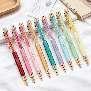 Peut inclure: Une collection de stylos personnalisés de différentes couleurs, dont le rouge, le rose, le jaune et le turquoise. Chaque stylo est orné d'un corps rempli de cristaux et d'éléments dorés. Des noms comme James, Emma et Daniel sont gravés.