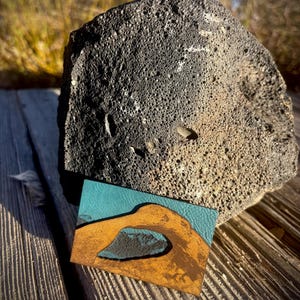 Könnte beinhalten: Ein rechteckiges Lederstück mit einem Landschaftsdesign, das einen türkisfarbenen Himmel und einen goldenen Torbogen zeigt. Das Stück liegt auf einer Holzoberfläche neben einem großen, strukturierten, dunkelgrauen Felsen.