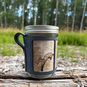 May include: A glass jar with a silver metal lid and a dark blue handle. The jar is wrapped in a dark blue sleeve featuring a central panel of light-colored birch bark with a dark brown, laser-engraved silhouette of a jumping fox.