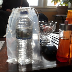 May include: A clear plastic bag with a drawstring closure contains a clear plastic bottle of Glacier Spring water. The bottle is sitting on a wooden table next to a clear glass bowl filled with colorful candies and two bottles of perfume.