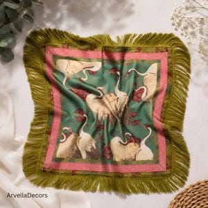 May include: A square scarf featuring a green background with a pattern of white cranes and red floral accents. The scarf is bordered by a pink frame and finished with long, olive green fringe along all four edges. The text ArvellaDecors is visible.