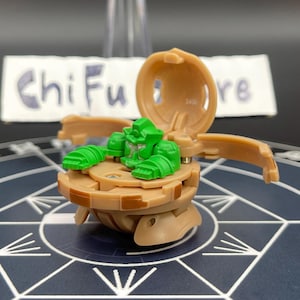 May include: A tan and green toy figure with a hinged shell, open to reveal a green character. The toy is on a patterned surface, with the text "Chi Fu" visible in the background.