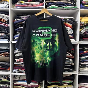 May include: Black t-shirt featuring the text "COMMAND & CONQUER" in white and green. The graphic includes a portrait of a man with green and yellow accents. The shirt is hanging on a wooden hanger.
