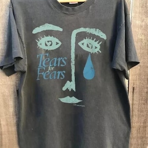 May include: Dark grey t-shirt with a blue face design and the words "Tears for Fears". The design includes eyes, a nose, and a teardrop. The t-shirt is hanging on a wooden hanger.