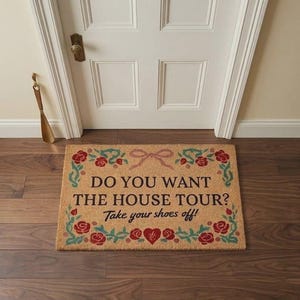 Op de afbeelding: Een rechthoekige deurmat met de tekst "DO YOU WANT THE HOUSE TOUR? Take your shoes off!" omgeven door rode rozen en groene bladeren. Bovenop zit een lichtbruine strik. De mat ligt op een houten vloer.
