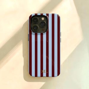 Op de afbeelding: Een smartphonehoesje met een verticaal streeppatroon in afwisselende tinten bordeauxrood en lichtblauw. Het hoesje heeft een uitsparing voor de camera linksboven en de goudkleurige initialen ML in een schreeflettertype onderaan in het midden.