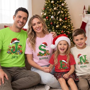 May include: A set of four t-shirts in lime green, light pink, red, and cream, each featuring a large letter graphic decorated with a Santa hat, Christmas gifts, and an elf, with the names Jack, Sophia, Emily, and Noah printed below.
