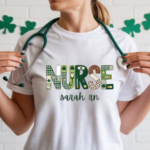 Puede incluir: Camiseta blanca con la palabra "NURSE" en un diseño de patchwork con cuadros verdes, estampado de leopardo y símbolos médicos. El nombre "sarah, rn" está impreso debajo. Un estetoscopio verde cuelga del cuello.