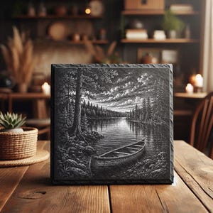 May include: A square slate plaque featuring a black and white engraved landscape illustration of a canoe resting on the rocky shore of a calm lake, surrounded by a dense pine forest under a cloudy sky with a textured, rough-hewn edge finish.