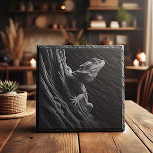 Könnte beinhalten: Eine quadratische, schwarze Schiefertafel zeigt ein detailliertes Relief einer Bartagame, die einen Baum erklimmt. Das Kunstwerk ist in Graustufen gehalten, wobei die Schuppen der Eidechse und die Rinde des Baumes detailliert dargestellt sind. Die Tafel steht auf einem Holztisch.
