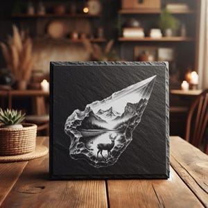 May include: A square, black slate coaster with a detailed, etched landscape scene featuring mountains, a lake, and a deer. The coaster is sitting on a wooden table.