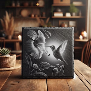 Peut inclure: Une dalle carrée en ardoise noire gravée d'un colibri et d'une fleur d'hibiscus. Le motif en niveaux de gris représente l'oiseau en vol près de la fleur. La dalle est posée sur une table en bois. Mesure environ 20 cm x 20 cm.