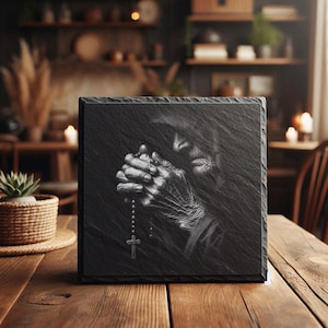May include: A square, black slate tile features a detailed grayscale image of a person's hands clasped in prayer, holding a rosary with a cross. The image is set against a dark background, creating a dramatic and reverent feel.