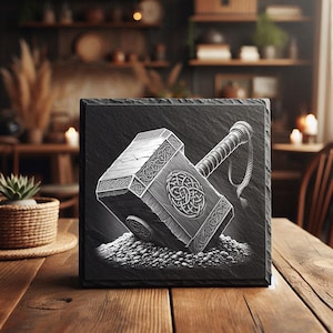 Puede incluir: Un posavasos cuadrado de pizarra negra con una ilustración detallada en escala de grises del martillo de Thor, Mjolnir. El martillo descansa sobre un montón de pequeñas piedras. El mango tiene un detalle de cuerda. El posavasos está sobre una mesa de madera.