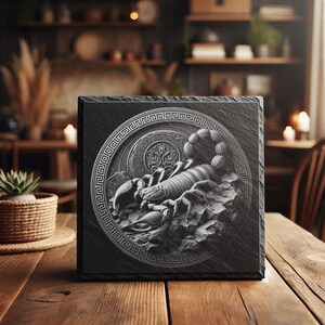 Puede incluir: Un posavasos cuadrado de pizarra gris oscuro con un diseño de escorpión grabado en detalle. El escorpión está centrado dentro de un borde circular ornamentado. El posavasos está sobre una superficie de madera, lo que sugiere un artículo decorativo.