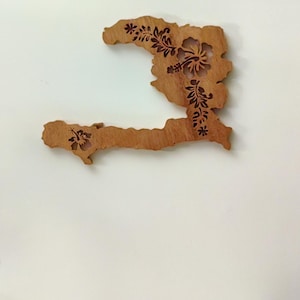 Peut inclure: Découpe en bois d'Haïti, ornée de motifs floraux. Le bois brun est façonné selon les contours du pays, avec des motifs d'hibiscus et autres motifs floraux découpés. La pièce est présentée sur un fond blanc.