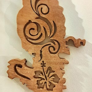 Può includere: Arte da parete in legno in una tonalità legno naturale, con un design intagliato di una forma stilizzata con motivi floreali e vorticosi. Il design include un fiore di ibisco e linee decorative.
