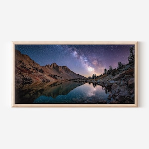 Könnte beinhalten: Ein gerahmtes Landschaftsfoto zeigt einen ruhigen See, der einen sternenklaren Nachthimmel und eine Bergkette widerspiegelt. Das Bild zeigt eine lebendige Darstellung der Milchstraße, wobei das Wasser die Himmelslandschaft widerspiegelt. Der Rahmen ist hellholzfarben.
