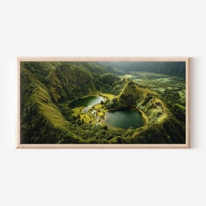 May include: A framed landscape print showcasing a vibrant green volcanic crater with two serene lakes. Lush vegetation covers the slopes, creating a natural, scenic view. The artwork is encased in a light-colored wooden frame.