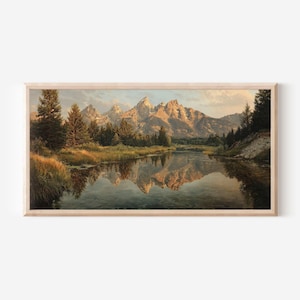 May include: A landscape painting in a light wood frame depicting a mountain range at sunrise with golden light hitting the peaks. A calm river flows in the foreground, reflecting the mountains and surrounding evergreen trees and tall grasses.