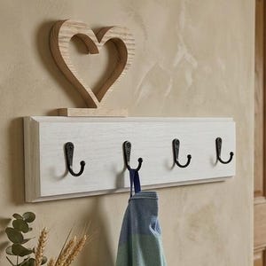 Peut inclure: Un porte-manteau en bois blanc avec quatre crochets noirs et une décoration en forme de cœur en bois. Une serviette à carreaux bleue et verte est suspendue à l'un des crochets. Une cruche en cuivre et une table en bois sont au premier plan.