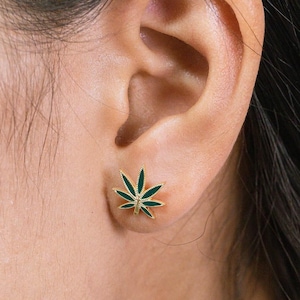 Puede incluir: Pendientes de botón dorados con forma de hoja de cannabis. El diseño de la hoja presenta esmalte verde oscuro que rellena los espacios entre las vetas doradas. Los pendientes son pequeños y se llevan en el lóbulo de la oreja.