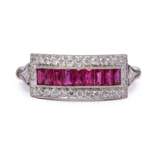 May include: A rectangular ring with a central row of vibrant red gemstones, framed by a border of smaller, clear stones. The ring has a silver-colored band and a detailed design on the sides, showcasing a classic jewelry style.