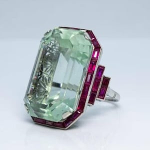 May include: A large, rectangular green gemstone ring with a silver band. The ring is accented with small, rectangular red gemstones around the green stone and on the side of the band. The ring is set against a white background.