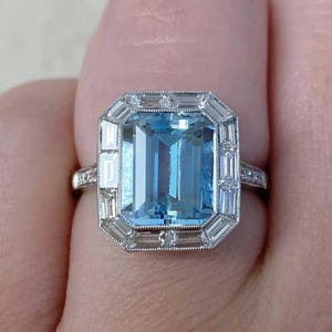 May include: A silver ring featuring a large, rectangular, light blue gemstone. The gemstone is surrounded by smaller, rectangular and round, clear gemstones. The ring is set on a silver band.