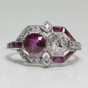 May include: A vintage-style ring featuring a large, round, deep-red ruby and a large, clear, round-cut diamond. The ring is set in a silver or white gold band, with smaller diamonds and red stones accenting the design. The ring has an Art Deco style.
