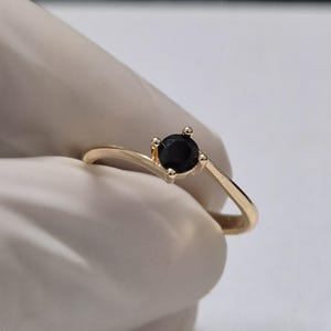 May include: A gold-colored ring with a single, round black gemstone. The ring has a delicate band and a simple, elegant design. The gemstone is set in a four-prong setting.