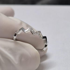 May include: A silver-colored ring with a mountain-like design. The ring features small, clear gemstones set along the peaks of the mountain range. The ring is held by a gloved hand against a white surface.