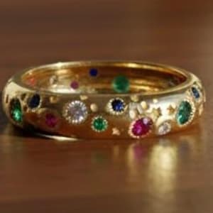 May include: A gold ring with a band adorned with various colorful gemstones, including green, red, blue, and white. The ring has a polished finish and is set on a wooden surface. The gemstones are set in a pattern of circles and stars.