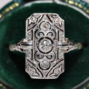 Puede incluir: Un anillo de estilo Art Déco con una cara rectangular, con un diamante central y detalles intrincados de filigrana. El anillo está engastado en un metal plateado y se muestra en un joyero de terciopelo verde.