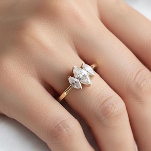 Puede incluir: Un anillo de oro con un grupo de diamantes talla marquesa. Los diamantes están dispuestos en un patrón floral, con tres piedras más grandes en el centro y otras más pequeñas a los lados. El anillo está engarzado en una fina banda de oro.