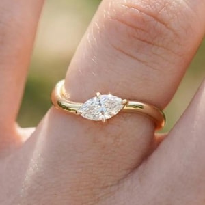 May include: A gold ring with a marquise-cut diamond. The ring has a twisted band design and is set with a clear, sparkling gemstone. The ring is shown on a finger against a blurred green background.