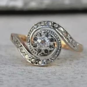 May include: A vintage-style ring with a central, round diamond surrounded by smaller stones. The ring features a swirling design with a gold band and a silver-toned setting for the diamonds. The ring is set against a neutral background.