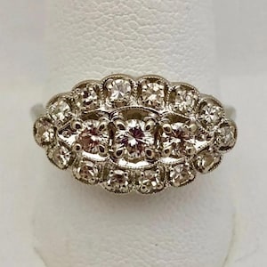 May include: A vintage-style ring with a cluster of diamonds. The ring features a central row of three larger diamonds, surrounded by smaller diamonds in a scalloped setting. The band is a light silver color.