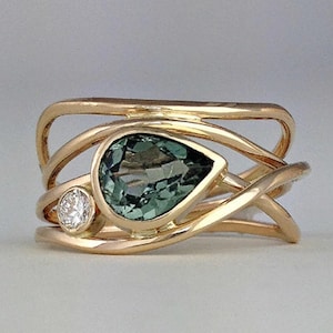 May include: A gold ring featuring a pear-shaped teal gemstone and a small round diamond. The ring has an intricate, interwoven design with multiple gold bands. The gemstones are set in a gold bezel.