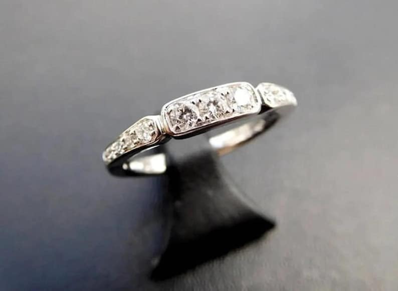 Puede incluir: Un anillo plateado con una fila de cinco diamantes redondos y transparentes engastados en un dise&ntilde;o rectangular. Se engastan diamantes m&aacute;s peque&ntilde;os adicionales en los lados del anillo. El anillo se muestra sobre un soporte negro sobre un fondo oscuro.