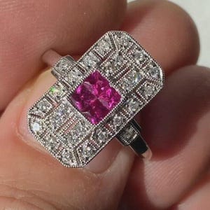 Peut inclure: Bague rectangulaire en argent avec une pierre précieuse rose vif carrée au centre. La bague est ornée de petits diamants transparents autour de la pierre rose et sur les bords du cadre rectangulaire.