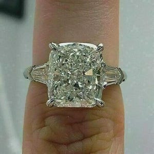 May include: A large, square-cut diamond ring with a brilliant, multifaceted center stone. The ring features smaller, rectangular diamonds on either side of the main stone, set in a silver or platinum band. The ring is a statement piece.