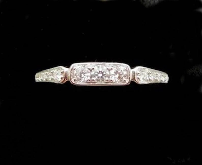 Puede incluir: Un anillo de plata con un engaste central rectangular con tres diamantes redondos. La banda se estrecha a cada lado, con diamantes m&aacute;s peque&ntilde;os engastados a lo largo de los hombros. El anillo est&aacute; sobre un fondo negro.