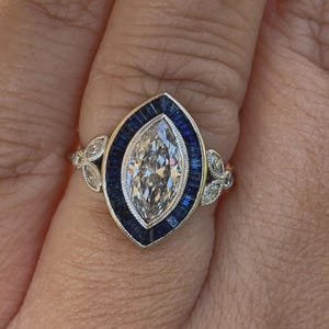 May include: A gold ring with a central marquise-cut diamond, framed by a border of blue sapphires. The ring showcases floral detailing on the shoulders, offering a classic, vintage aesthetic. The design is elegant and refined.