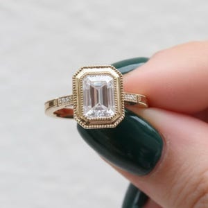 May include: A gold ring featuring an emerald-cut diamond center stone. The ring has a detailed bezel setting and small accent diamonds on the band. The ring is held by a person with dark green nail polish.