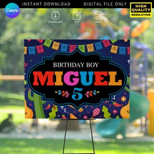 May include: A colorful birthday sign featuring the name "MIGUEL" in vibrant letters and the number "5". The design includes festive decorations like papel picado banners, cacti, and guitars, set against a dark blue background. The sign is displayed on a black easel.